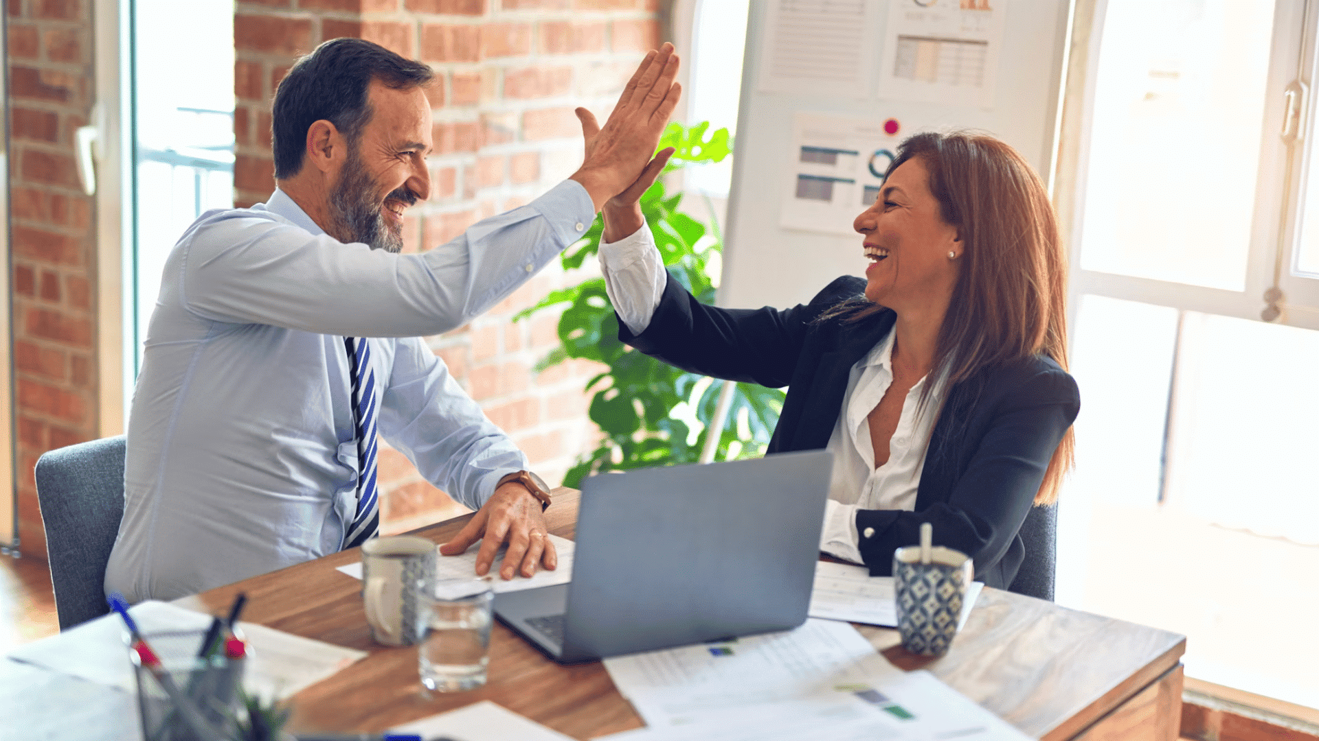 two coworkers smile and high five across a desk celebrating teamwork and a successful achievement