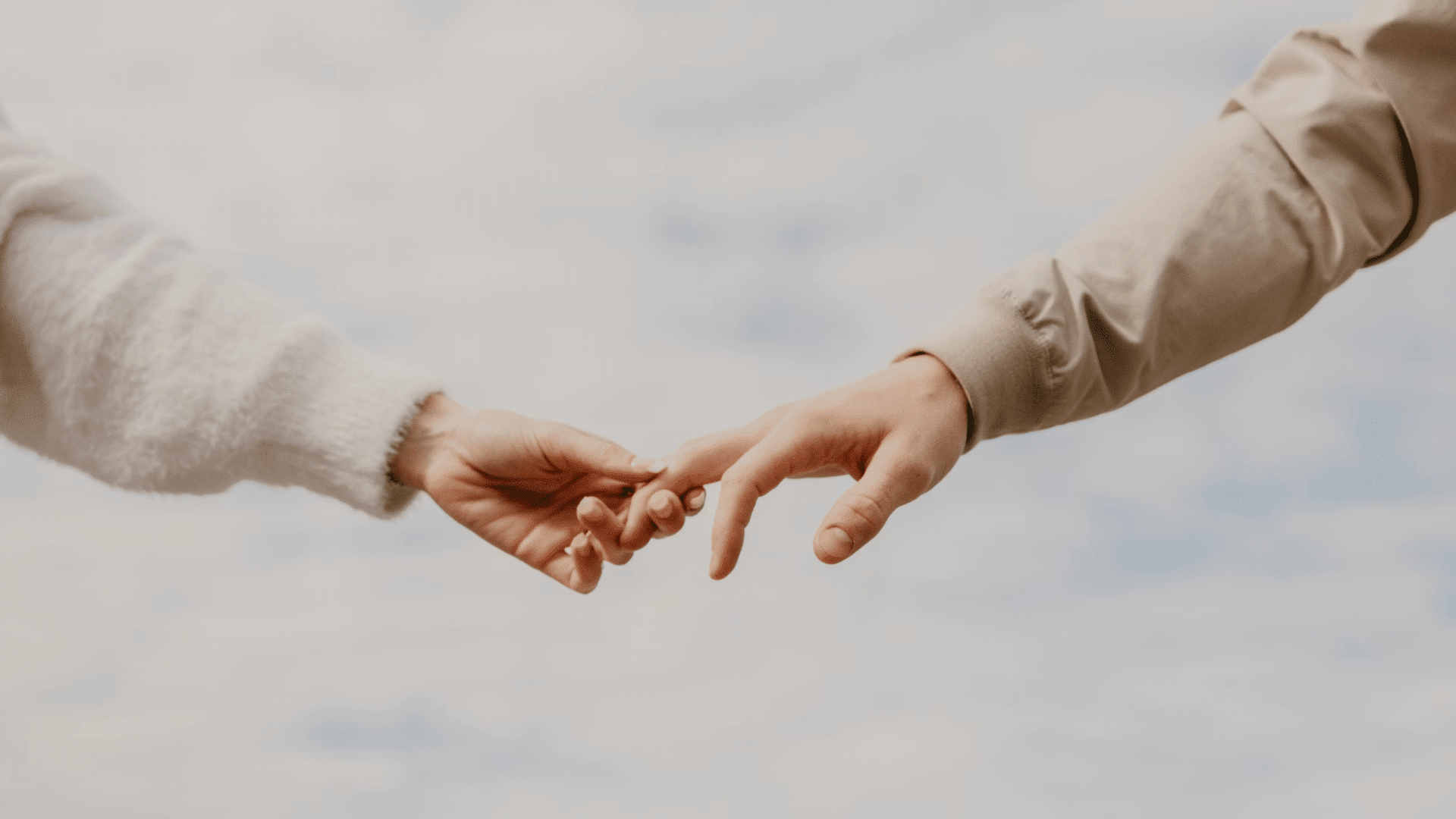 two people gently touching hands against a soft cloudy sky with pale sleeves framing the quiet moment