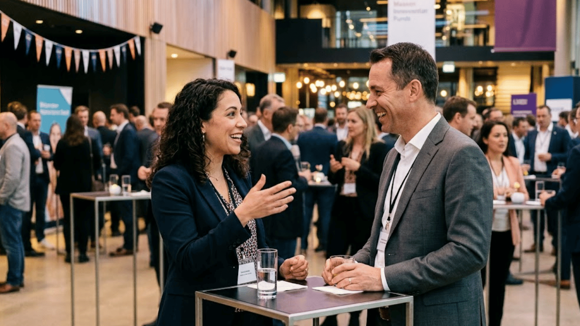 two professionals smiling while talking across a standing table at a busy networking event with attendees behind