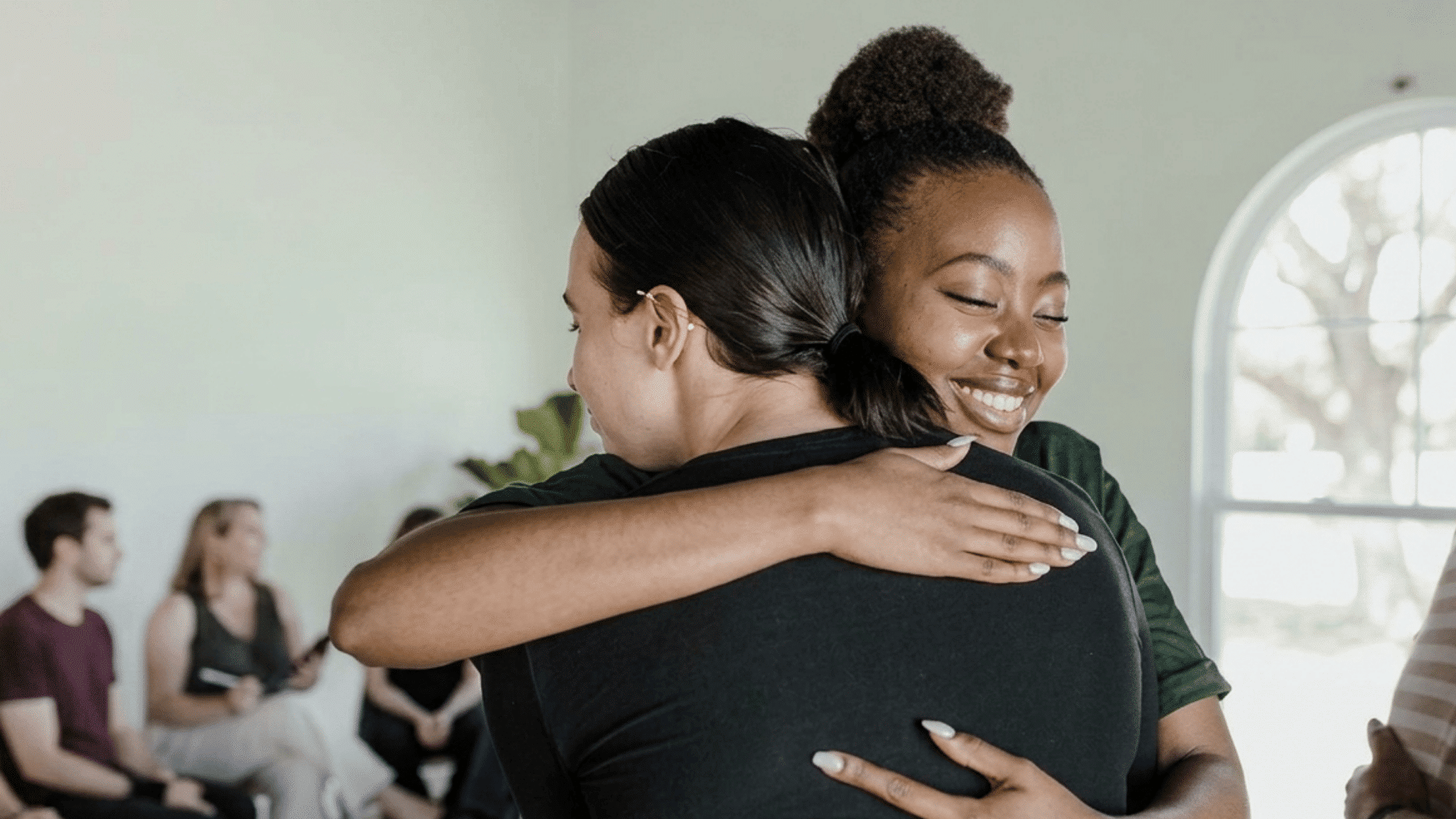 two women hugging during a support group meeting while others sit and talk in the background