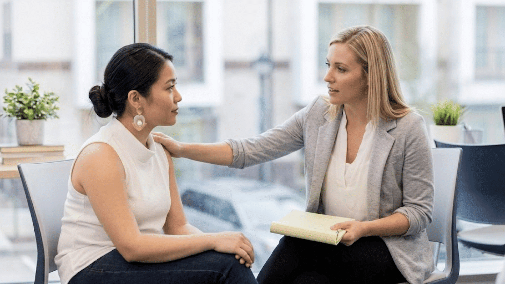 two women seated in an office having a supportive conversation with one woman gently touching the others shoulder