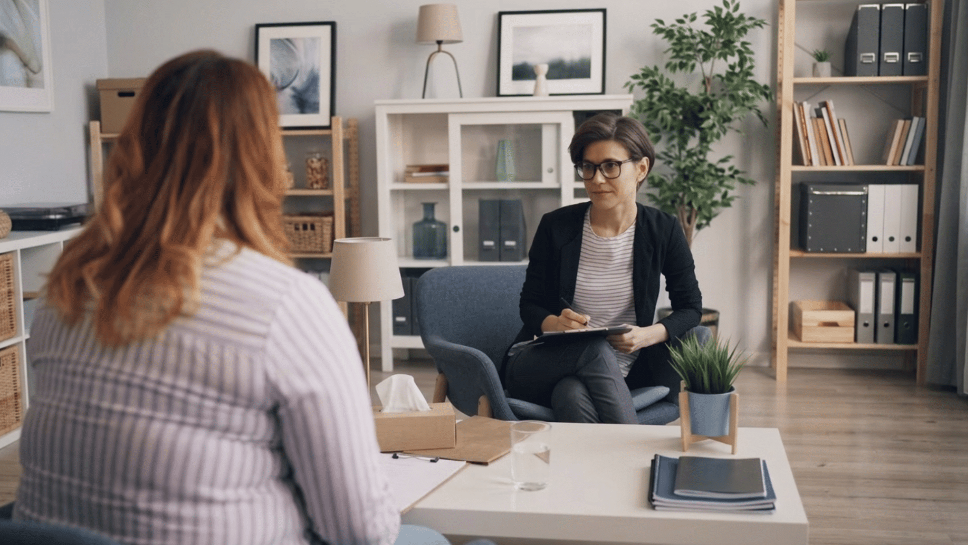 wide view of a therapist taking notes in an office opposite a patient in a striped shirt