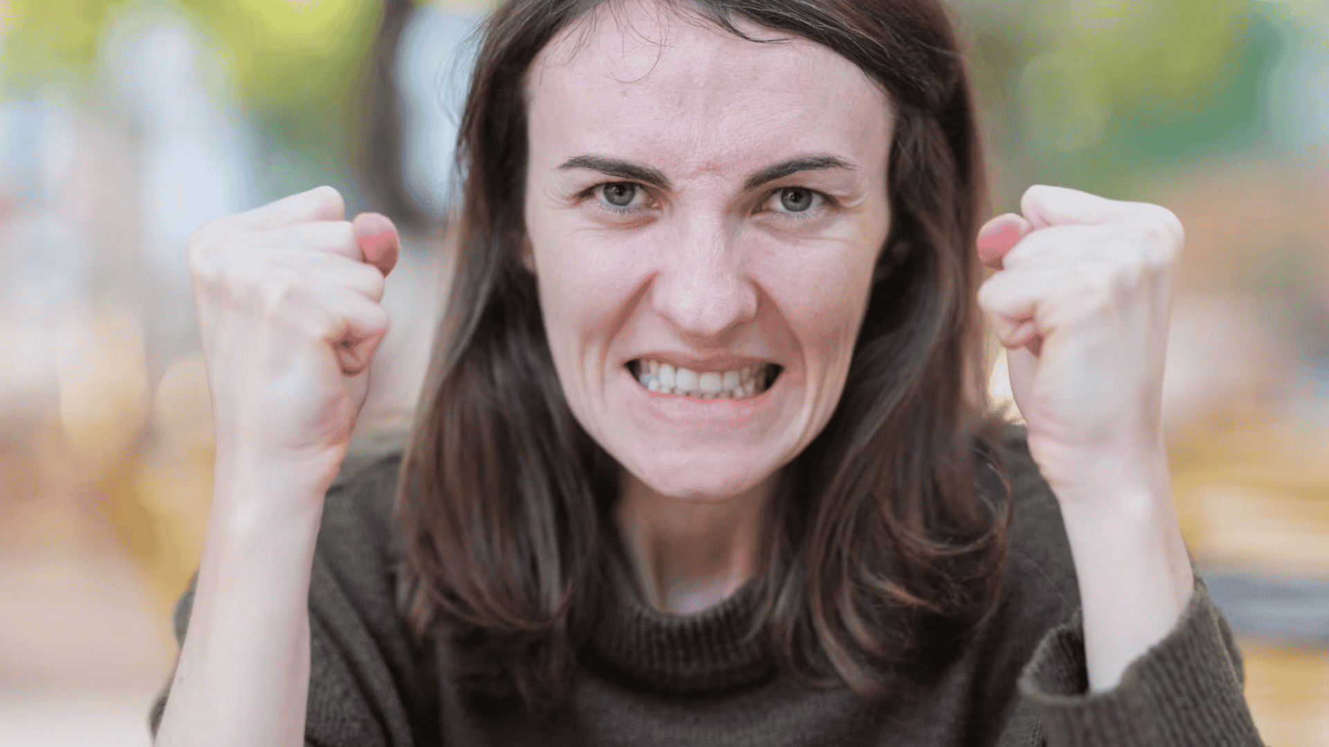 woman clenching both fists and showing an angry expression while looking directly at the camera outdoors