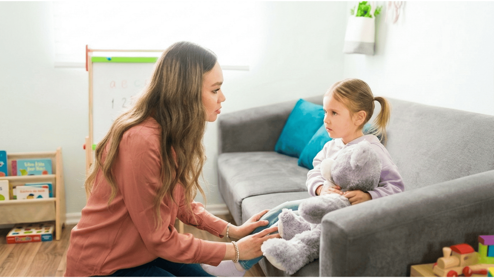 woman kneeling beside a young girl holding a teddy bear on a sofa