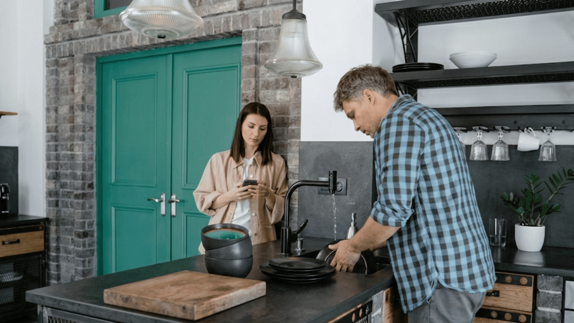woman looks at phone while man washes dishes in modern kitchen with teal doors