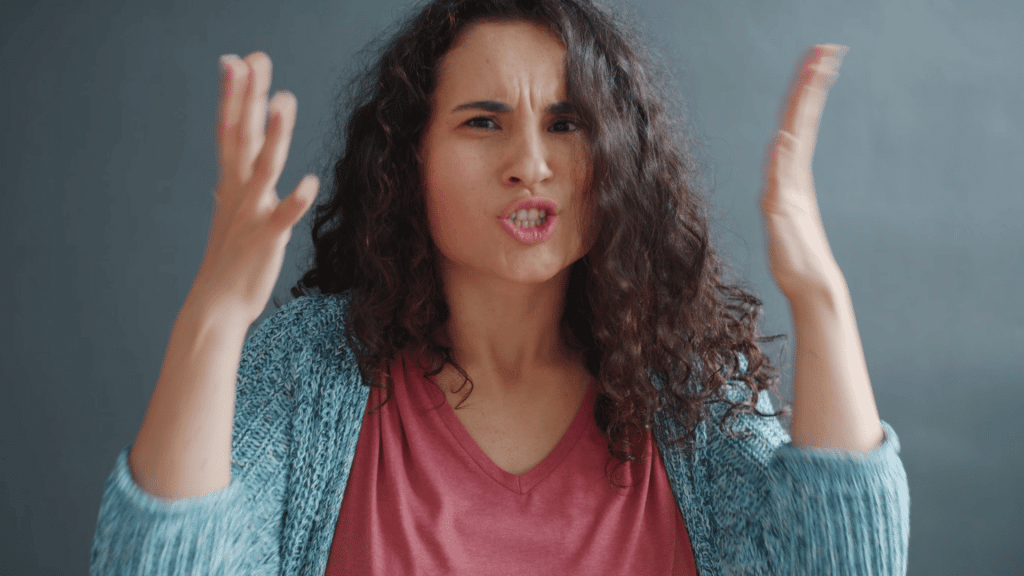 woman raising her hands with a frustrated expression while speaking against a plain indoor background
