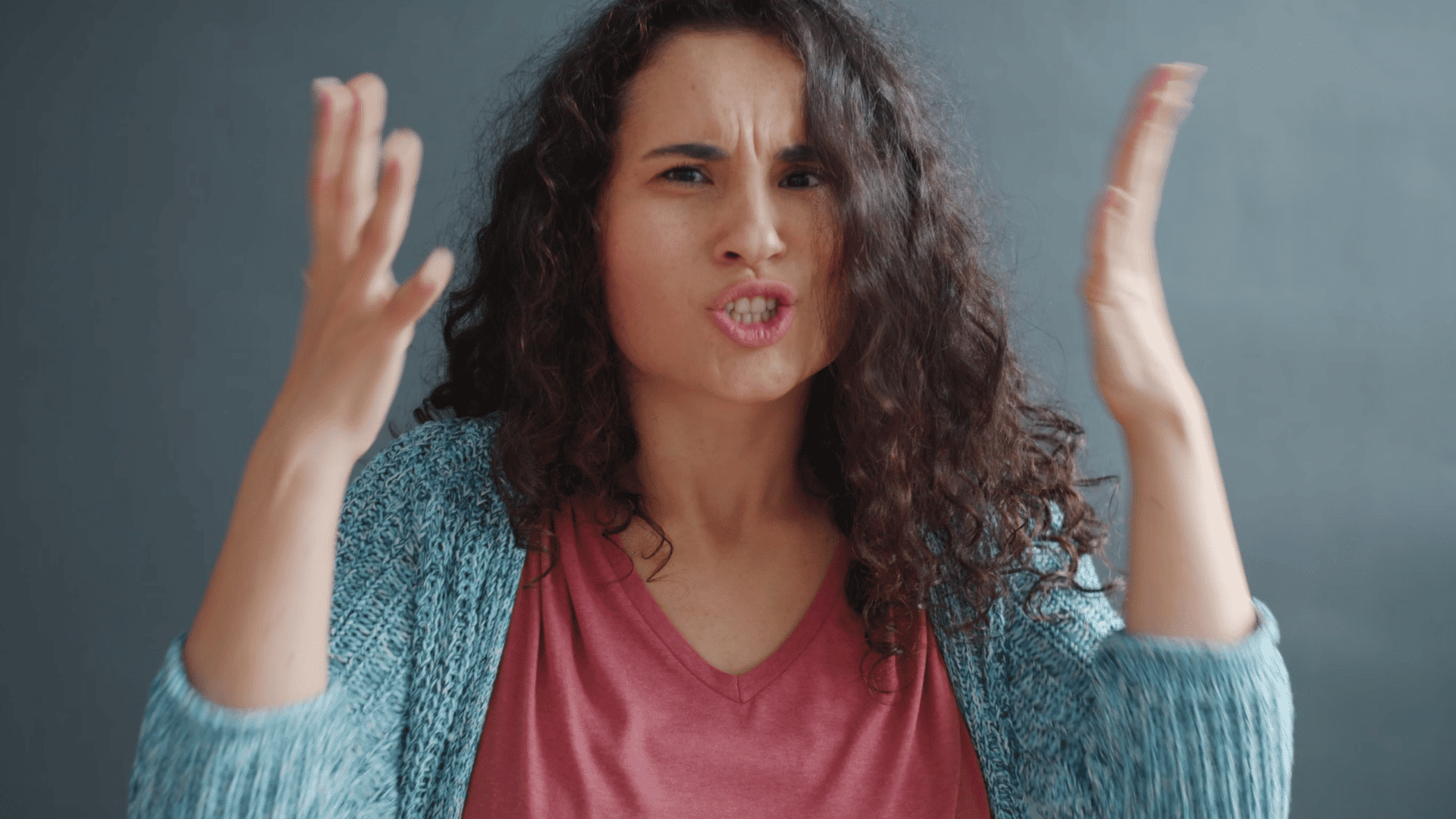 woman raising her hands with a frustrated expression while speaking against a plain indoor background