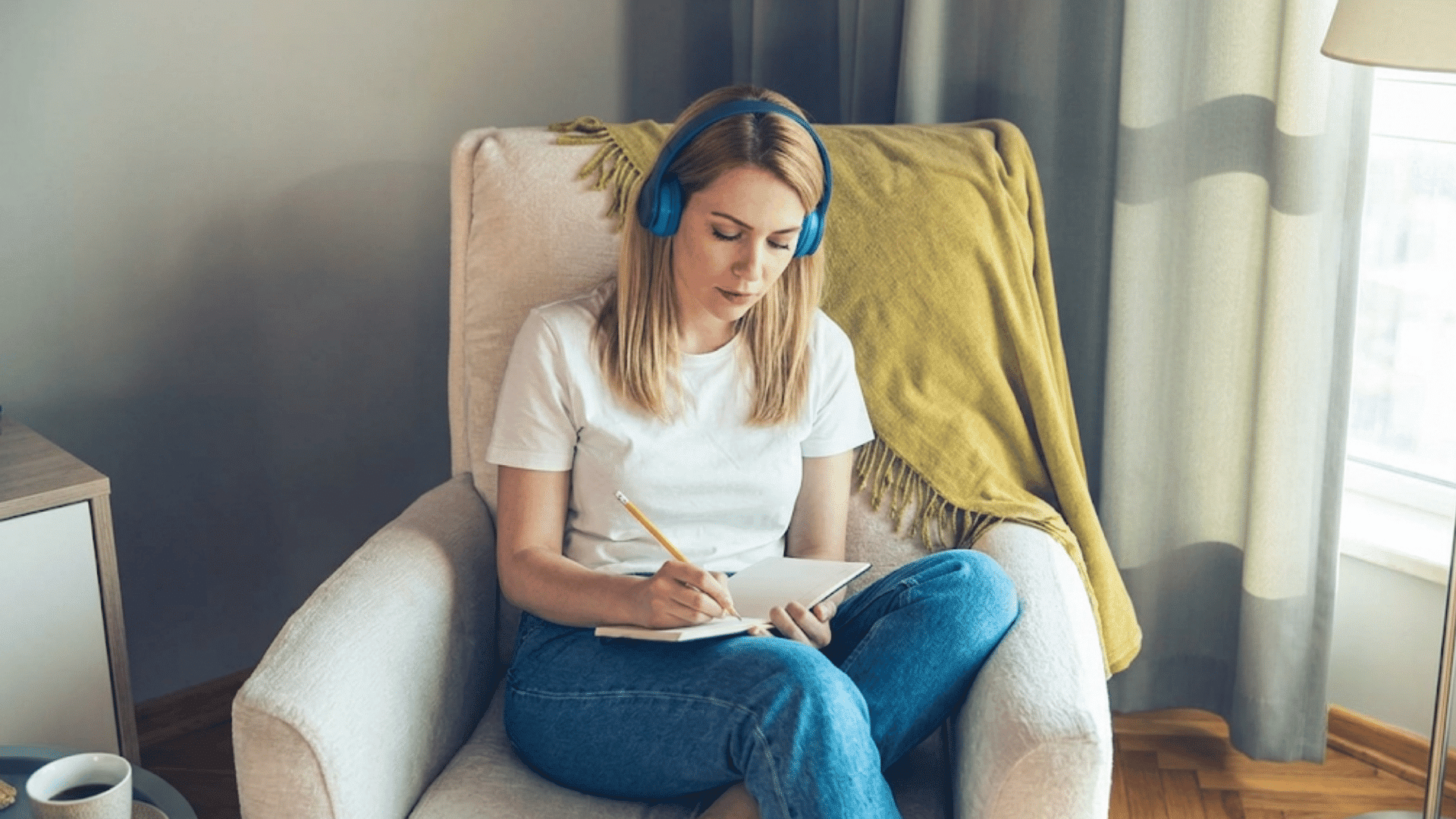 woman wearing blue headphones writes in a notebook while sitting in a chair near a bright window