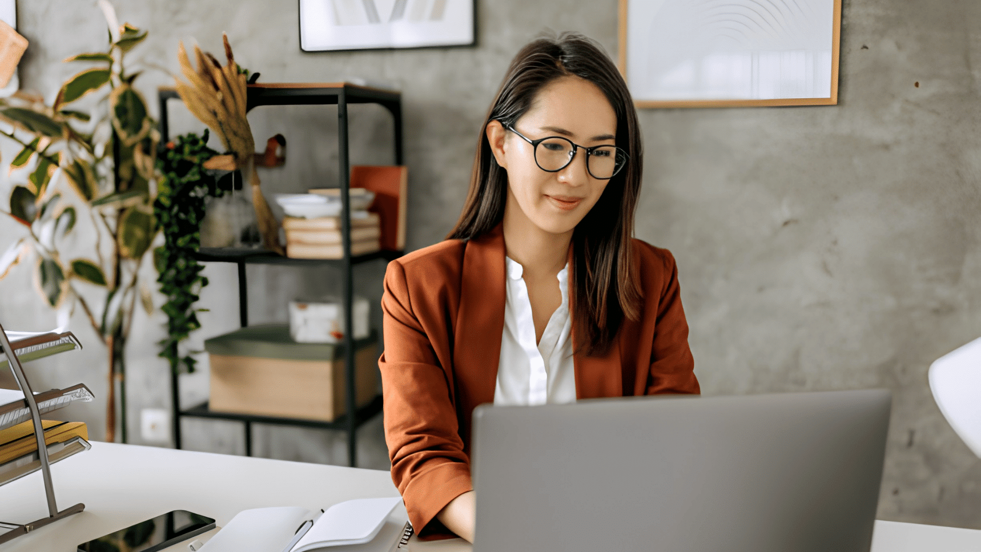 woman wearing glasses and a blazer working on a laptop in a modern office