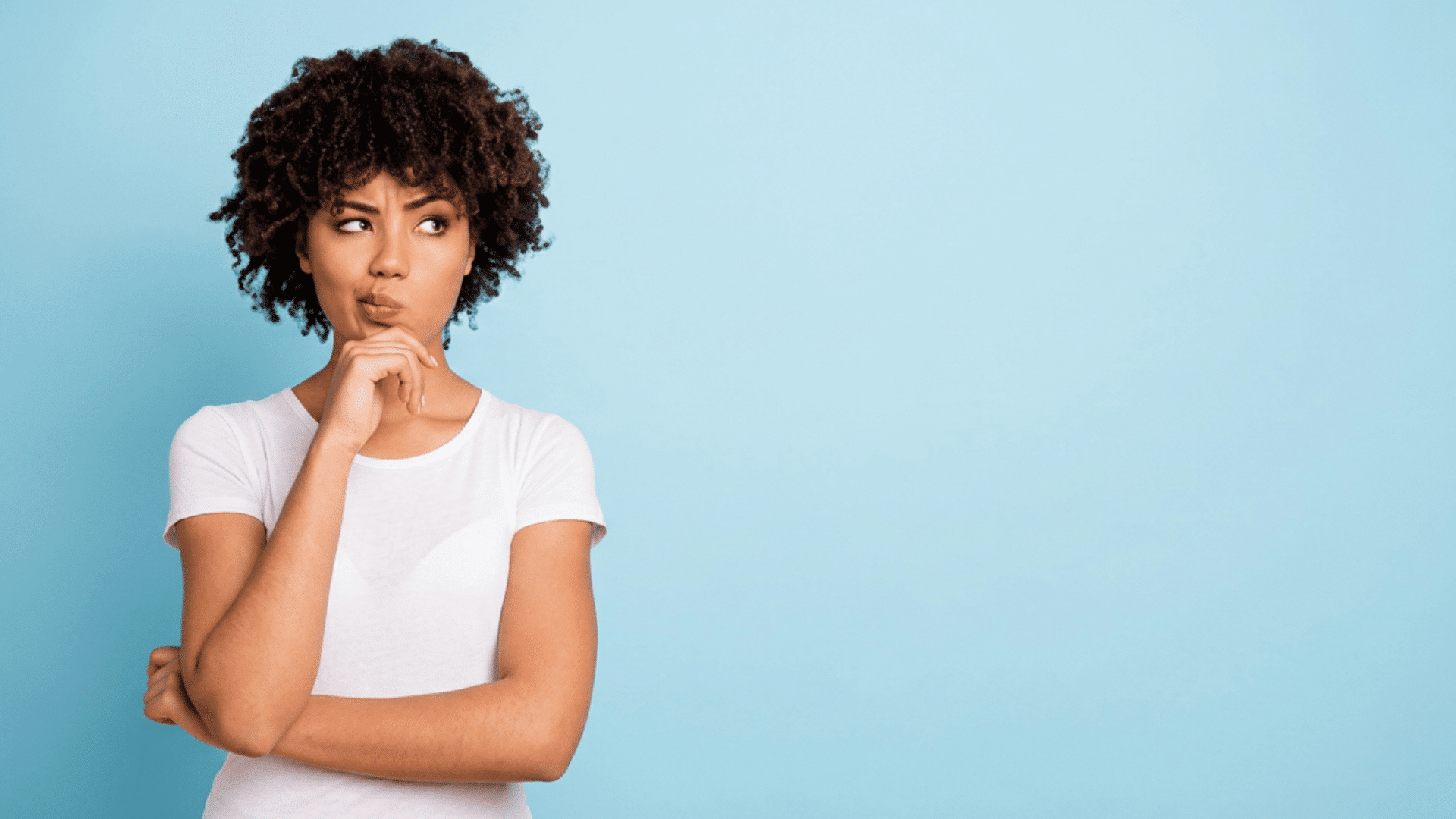 woman with curly hair in white shirt resting hand on chin and looking aside thoughtfully against blue background