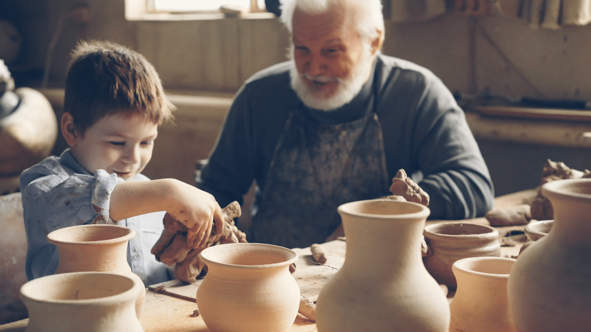young boy shaping clay beside an older potter surrounded by handmade bowls and vases in a warm workshop