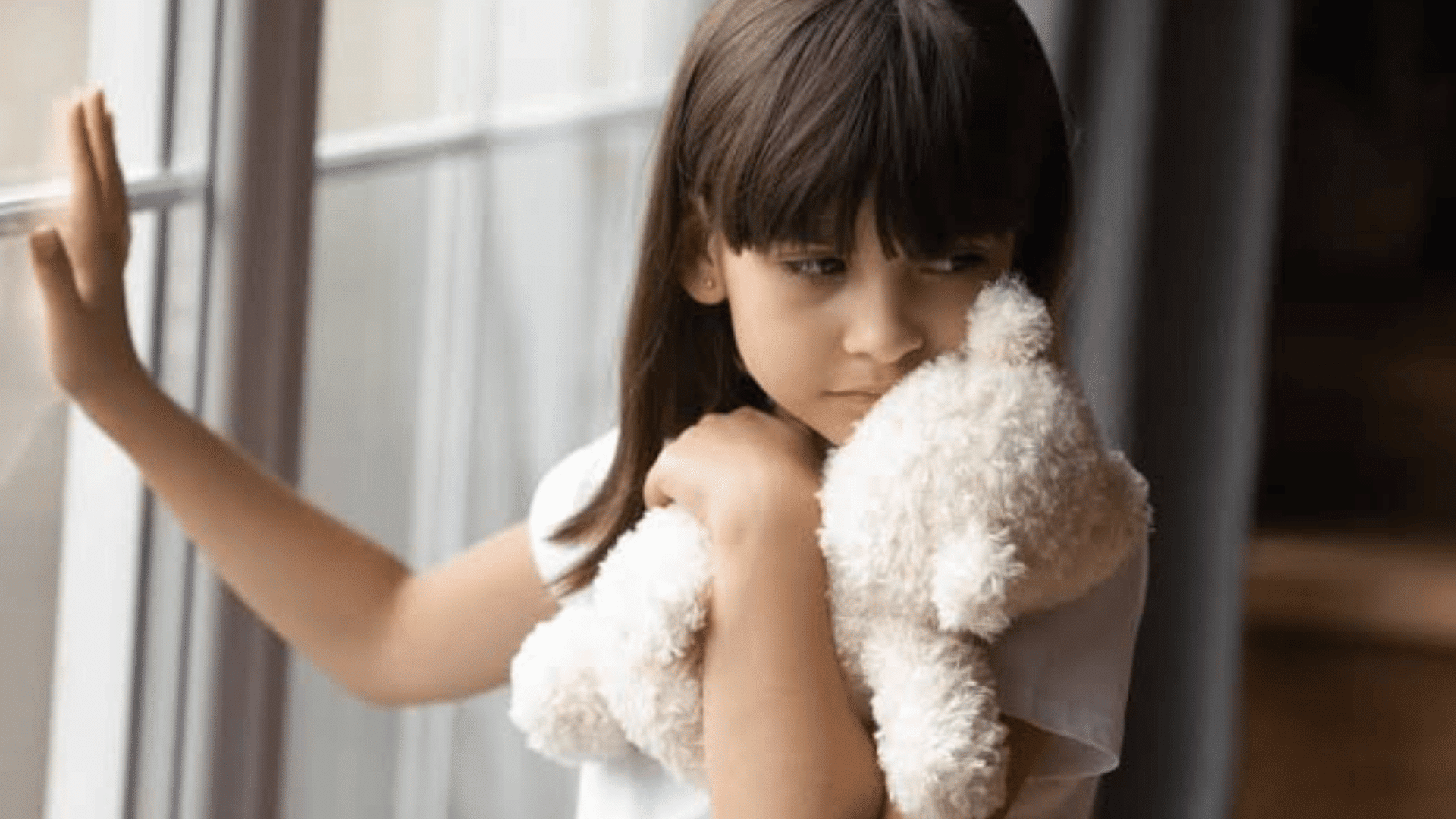 young child holding a stuffed toy while standing by a window with a sad and quiet expression