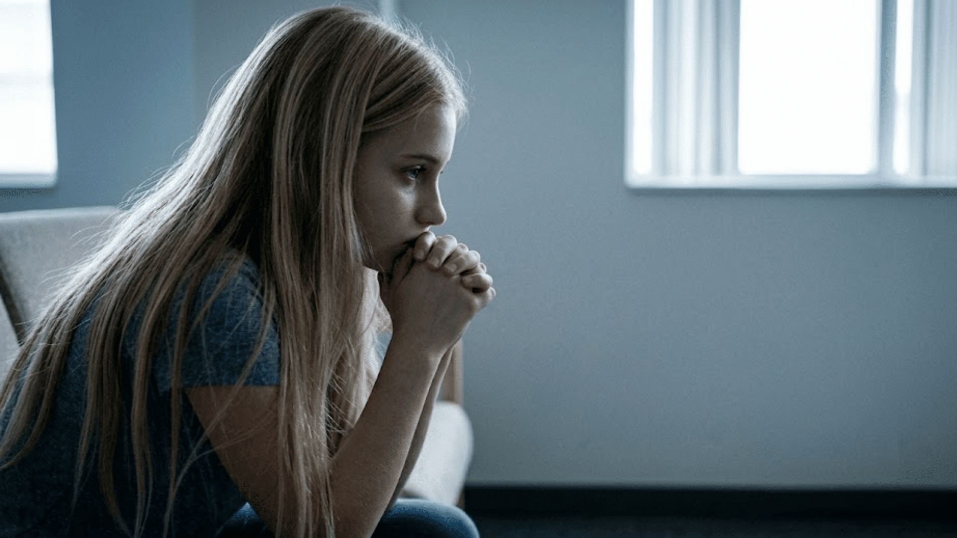 young girl sitting forward with hands clasped near her face looking thoughtful in a softly lit room indoors