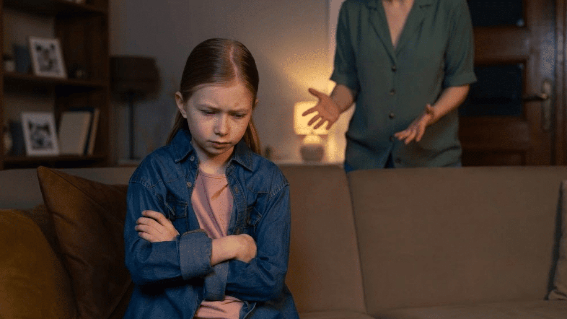 young girl sitting on a sofa with folded arms looking upset while an adult stands behind her gesturing during a tense moment at home
