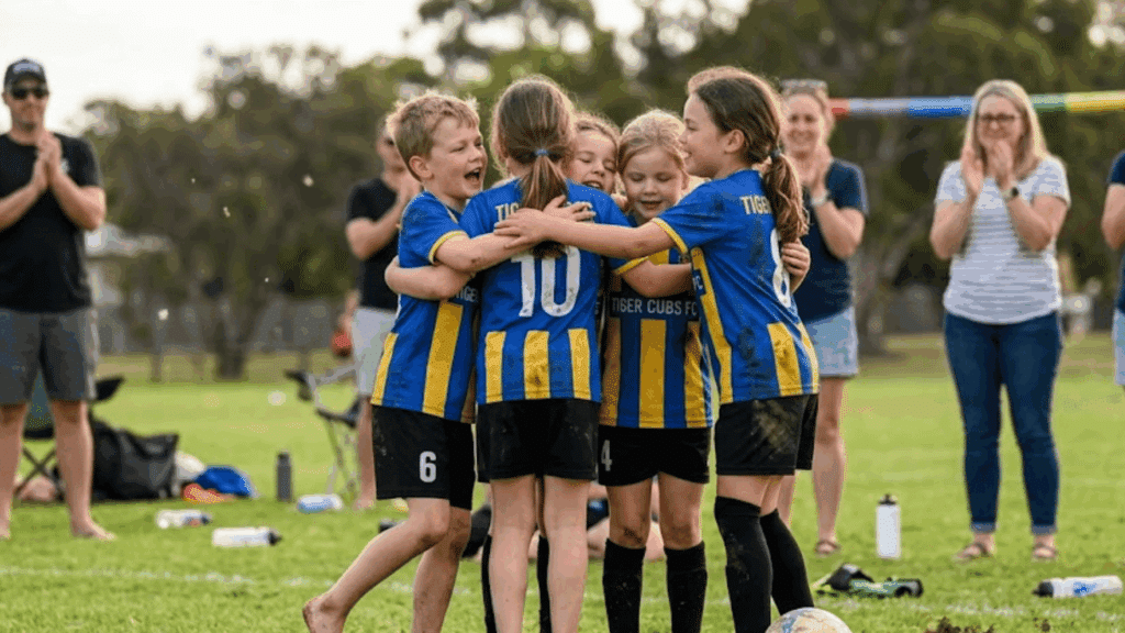 young soccer teammates wearing blue and yellow uniforms hugging in celebration on the field while spectators applaud in the background
