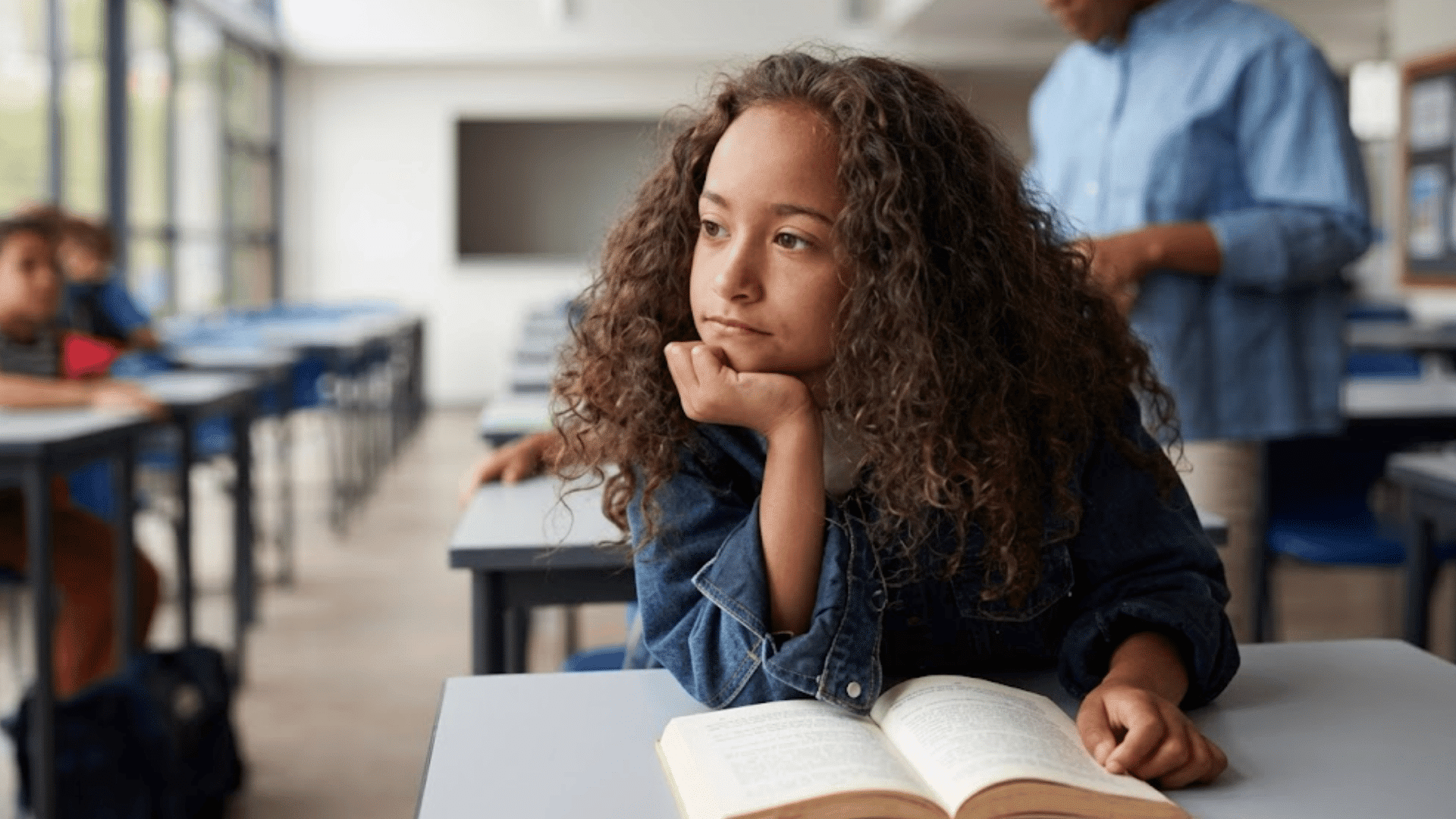 young student resting chin on hand while looking across classroom with open book on desk and classmates in background