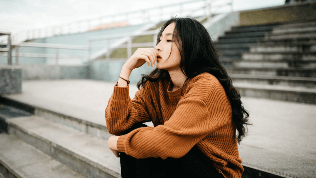 young woman sits alone on outdoor steps resting her hand near her face while gazing thoughtfully into the distance