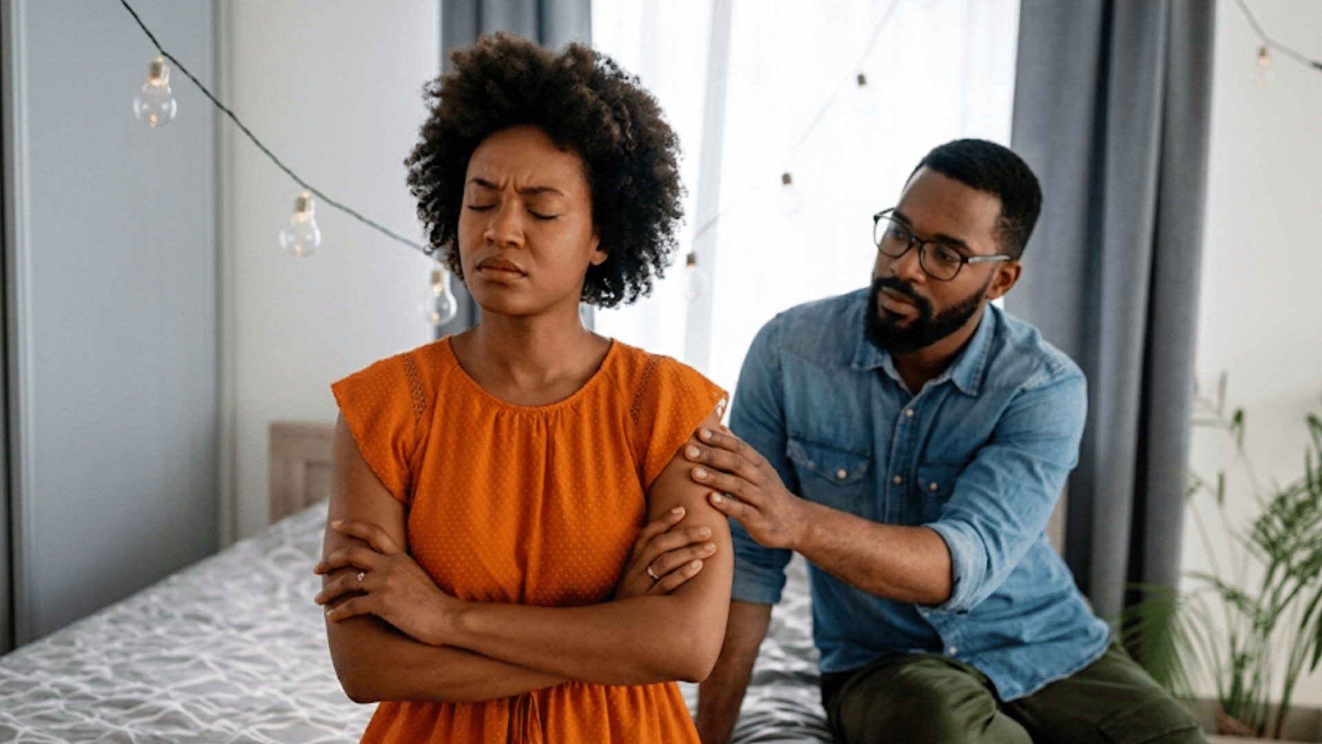 a woman sitting on a bed with folded arms while a man gently reaches toward her during a tense relationship moment