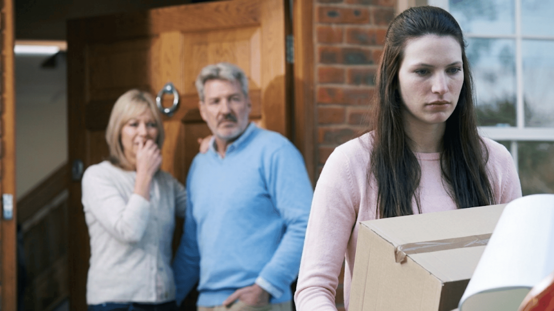 a young woman carrying moving boxes to a car while two older adults watch from the doorway with concerned expressions