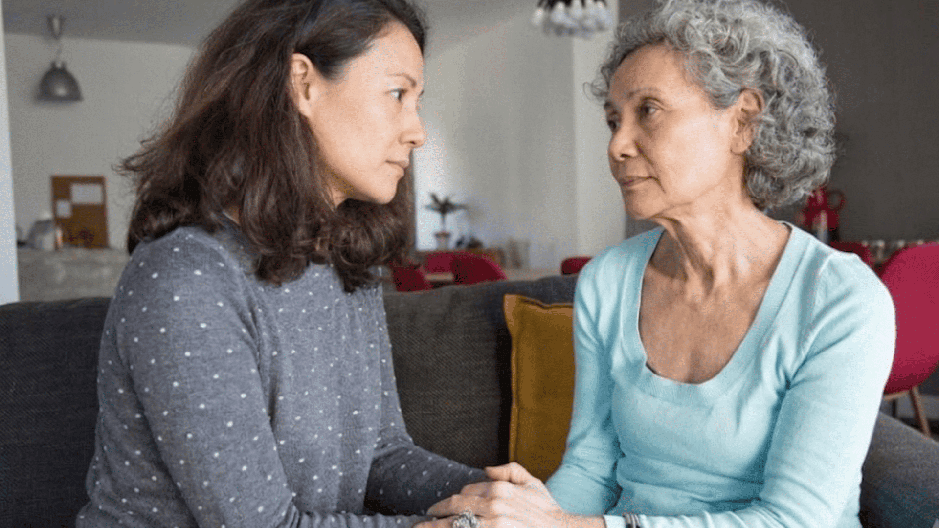 a younger woman holding an older womans hands as they share a caring conversation on a living room sofa