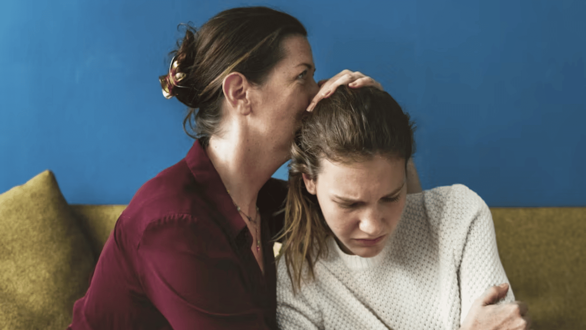 adult comforting a distressed young woman with a supportive embrace while they sit together on a sofa indoors