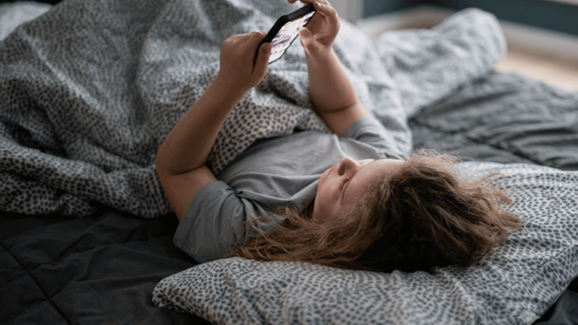 person lying on a bed holding a smartphone while relaxing under a blanket in a dim bedroom