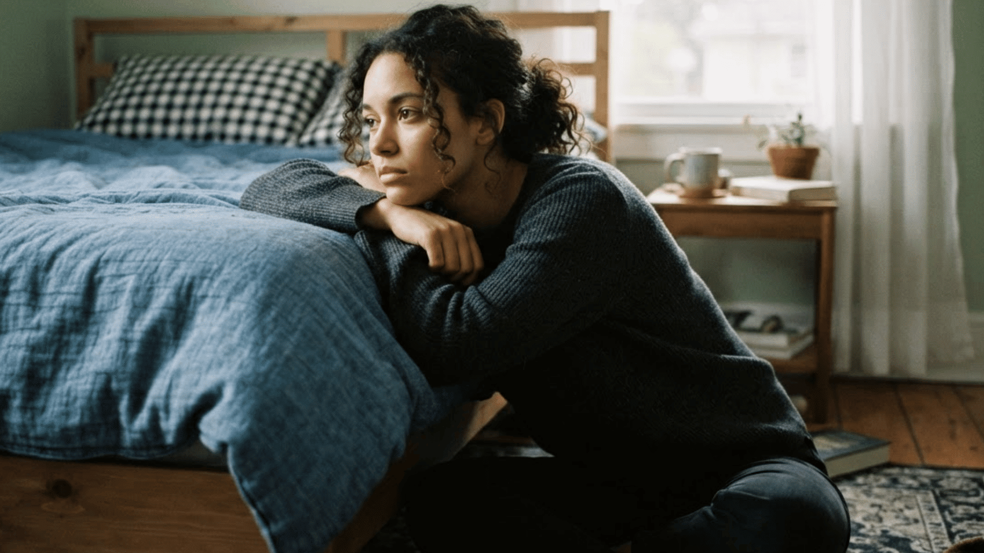 woman sitting on the floor beside a bed resting her arms and gazing out the window with a thoughtful expression