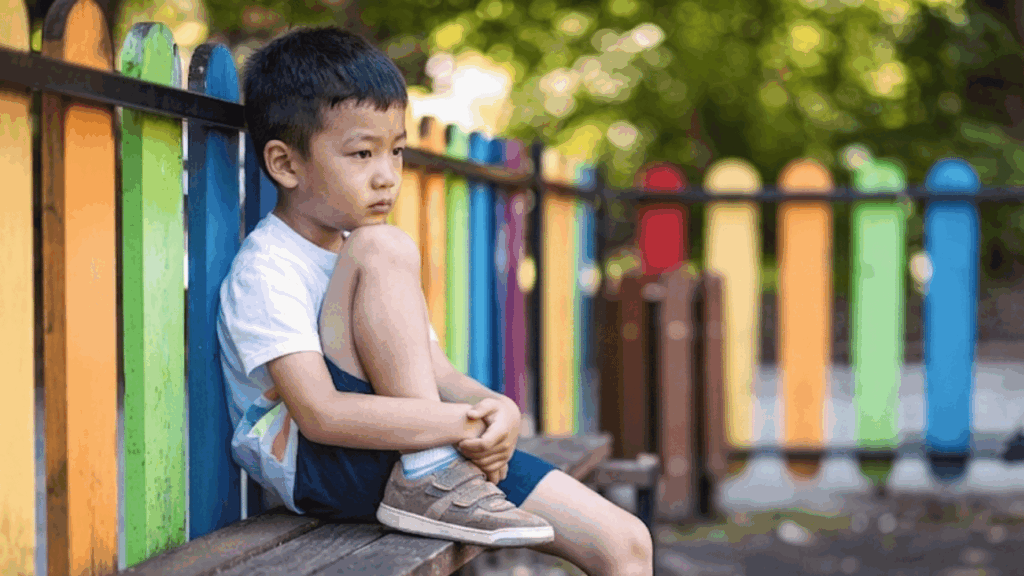 young boy sitting alone on a bench in a playground looking away with a thoughtful expression
