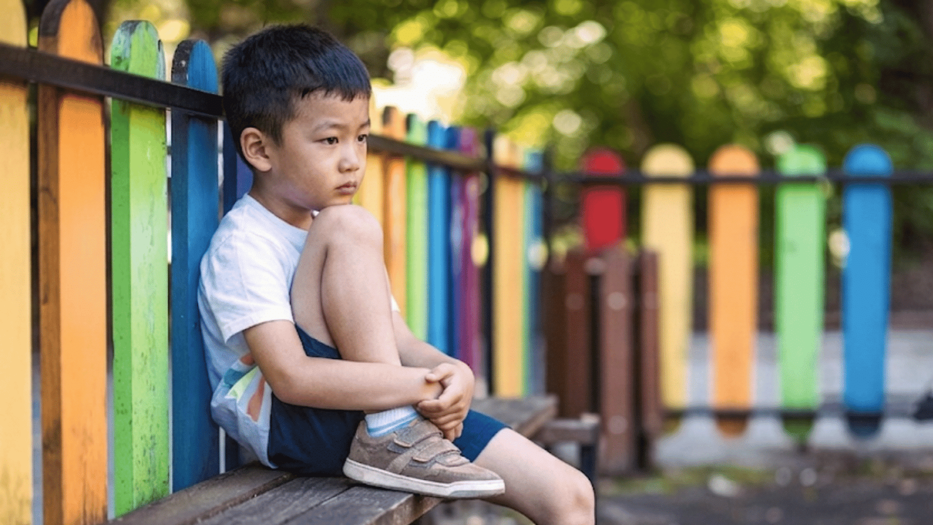 young boy sitting alone on a bench in a playground looking away with a thoughtful expression