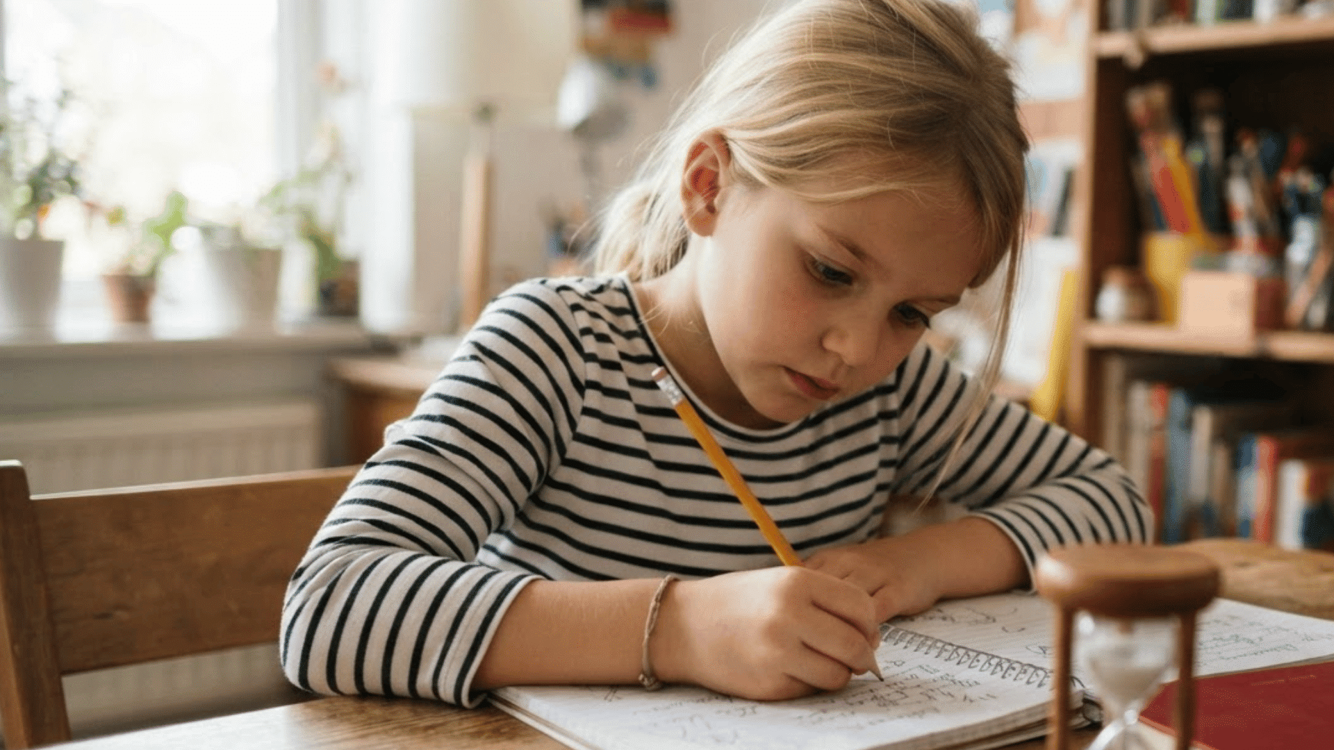 young child sitting at a table writing in a workbook with a pencil while focusing on schoolwork at home