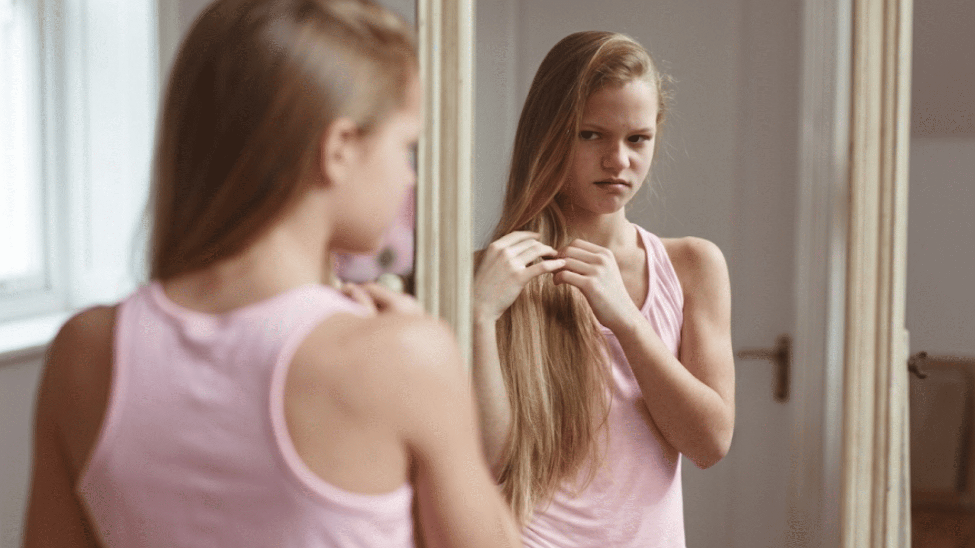 young girl looking at her reflection in a mirror while adjusting her hair in a bedroom setting